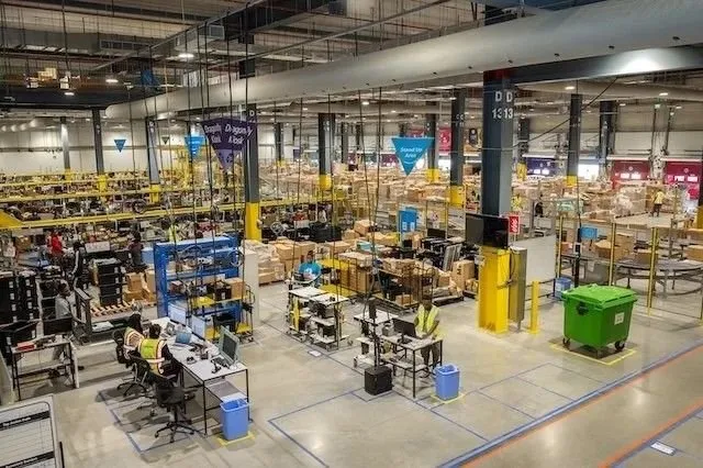 Busy Amazon warehouse interior with workers processing orders. Boxes and workstations fill the large space.