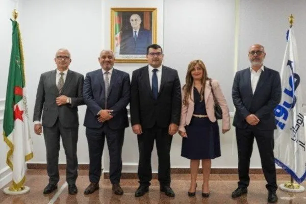 Five professionals in formal attire stand together, with Algerian flags and a portrait in the background.