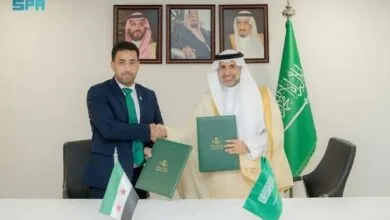 Two men shake hands at a signing ceremony, with flags and portraits of Saudi Arabian royalty in the background.