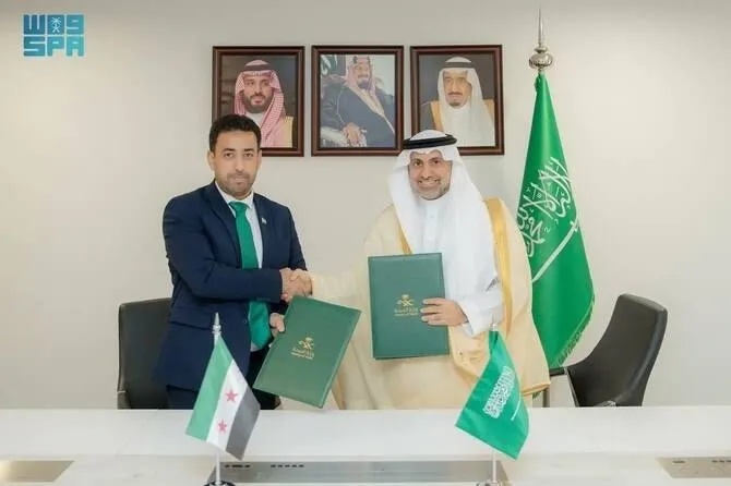 Two men shake hands at a signing ceremony, with flags and portraits of Saudi Arabian royalty in the background.