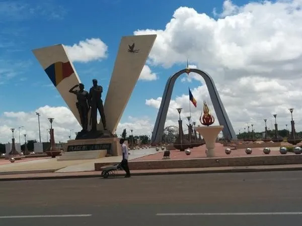 Monumental sculptures and arch in a plaza, under a bright blue sky with fluffy clouds. A person walks by with a wheelbarrow.