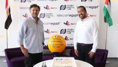 Two men smile at a launch event with a cake and balloon featuring logos for Coral Bridge Aqaba Landing, Telecomegypt, and Aqaba Digital Hub.
