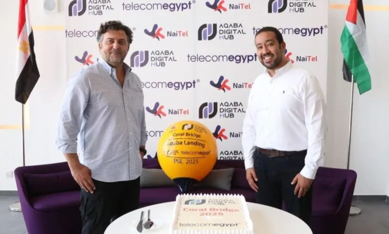 Two men smile at a launch event with a cake and balloon featuring logos for Coral Bridge Aqaba Landing, Telecomegypt, and Aqaba Digital Hub.
