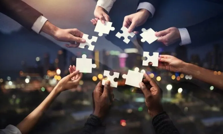 Diverse hands fitting together puzzle pieces against a city backdrop, symbolizing teamwork and collaboration.