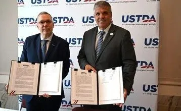 Two men in suits hold signed documents in front of a USTDA backdrop.