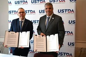 Two men in suits hold signed documents in front of a USTDA backdrop.