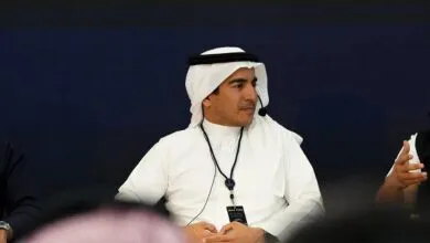 Man in traditional Saudi attire speaking at a conference, wearing a lanyard with an event badge.