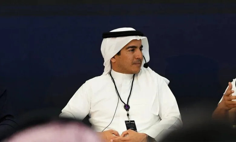 Man in traditional Saudi attire speaking at a conference, wearing a lanyard with an event badge.