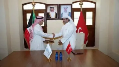 Two men in traditional Arab attire shaking hands in an office with flags and portraits.