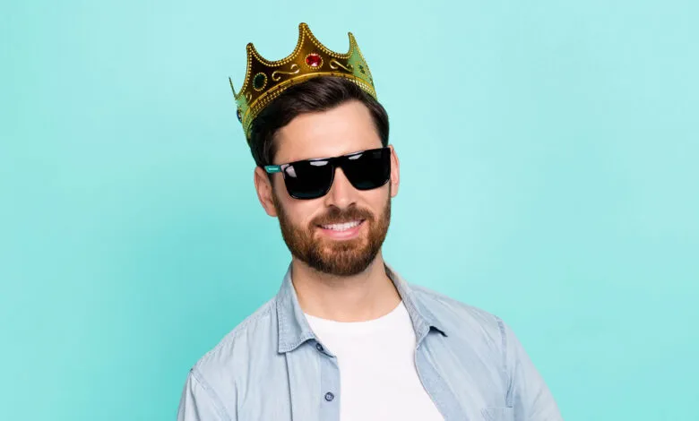 Smiling man wearing sunglasses and a gold crown against a light teal background.