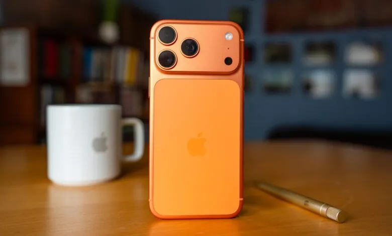 Orange iPhone 17 Pro Max on a wooden table with a pen and Apple mug.
