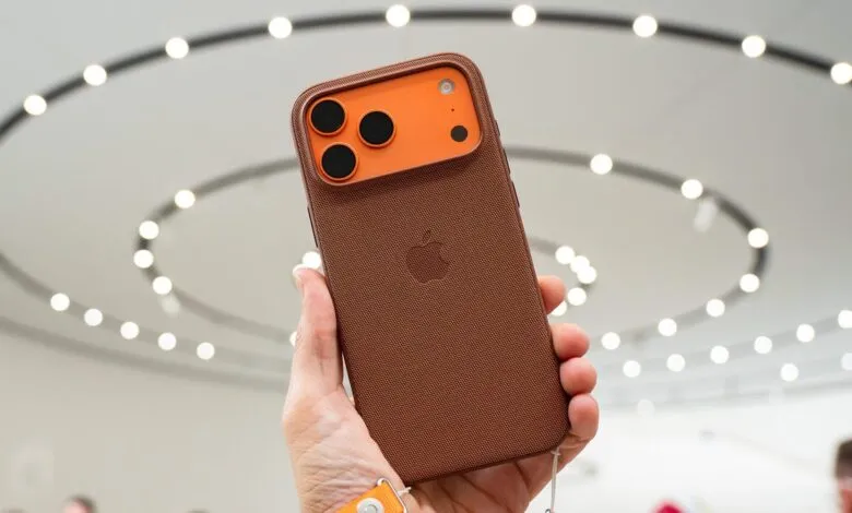 Person holding a brown iPhone 17 Pro Max with an orange camera housing. The background is blurred.