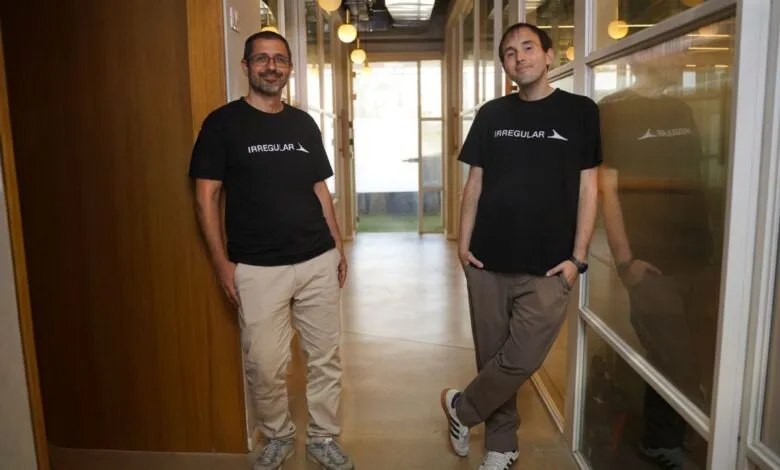 Two men in black Irregular Labs t-shirts stand in a modern office hallway.