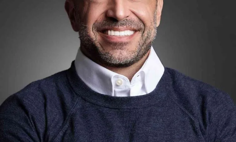 Headshot of Justin Kappers, smiling and arms crossed, wearing a navy sweater over a white shirt against a gray background.