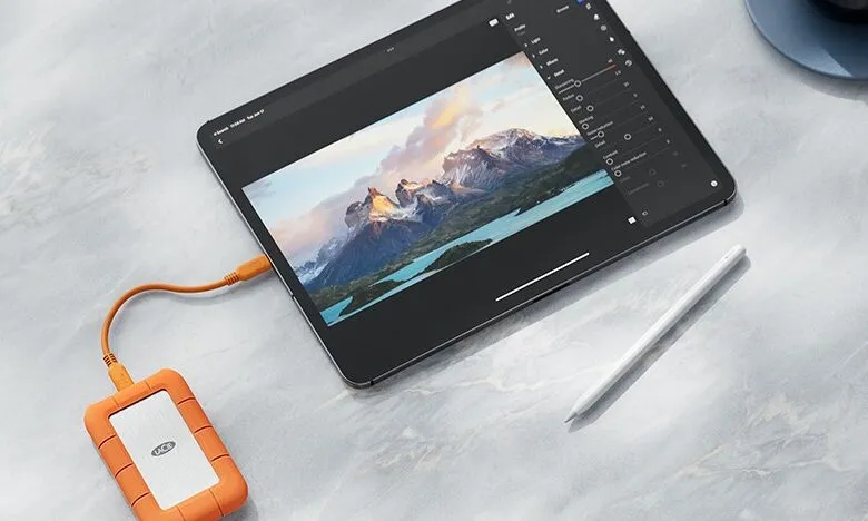 Tablet editing a landscape photo, connected to an orange LaCie hard drive with a stylus nearby.