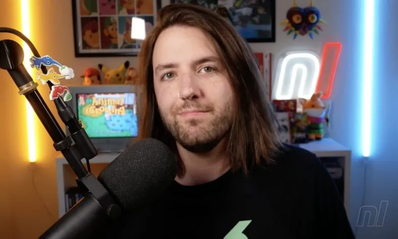 A man with long brown hair and a nose ring, wearing a black t-shirt, looks at the camera in a room with gaming memorabilia.