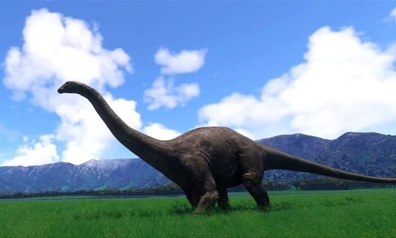 A long-necked dinosaur stands in a lush green field against a backdrop of mountains and a blue sky with fluffy white clouds.