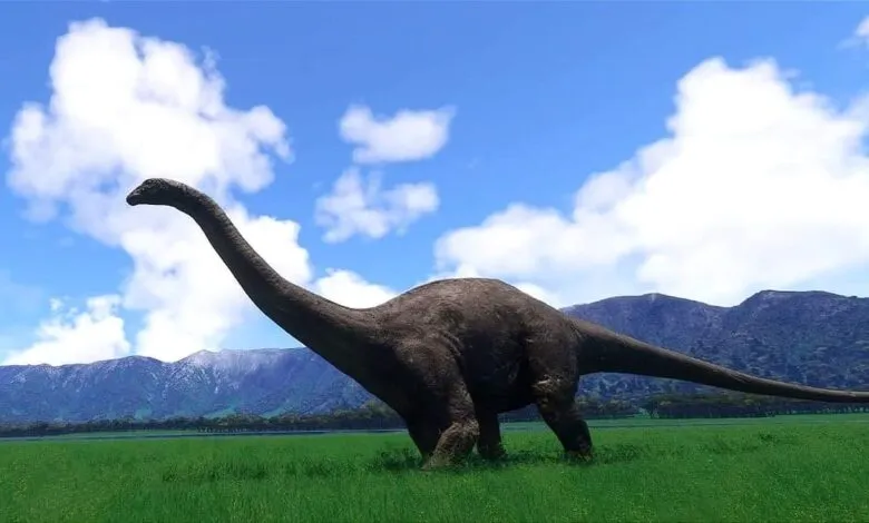 A long-necked dinosaur stands in a lush green field against a backdrop of mountains and a blue sky with fluffy white clouds.