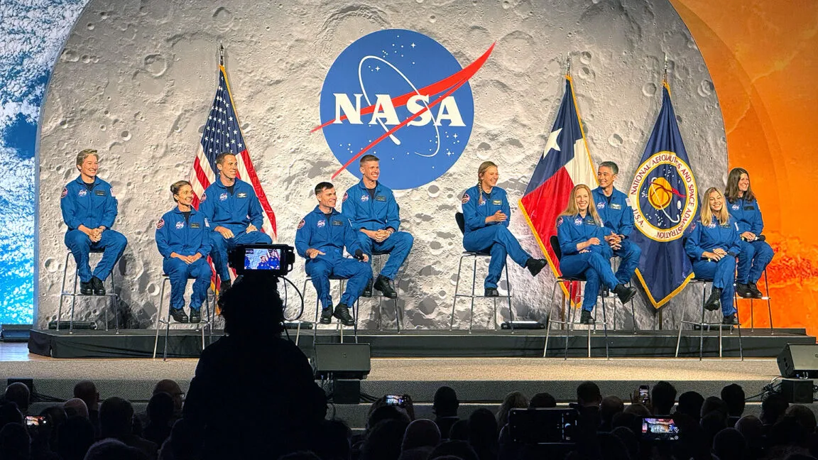 Artemis II astronaut crew seated on stage with NASA and Texas flags, against a moon backdrop.