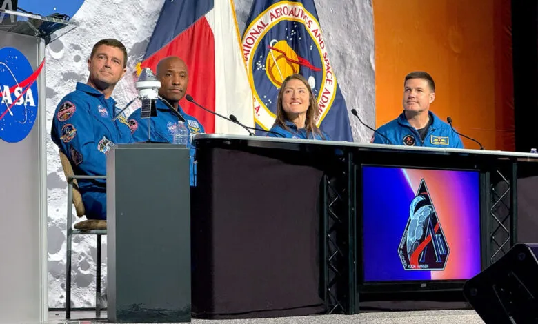 Four astronauts in blue suits sit at a table, NASA logo visible. A moon backdrop and flags are behind them.