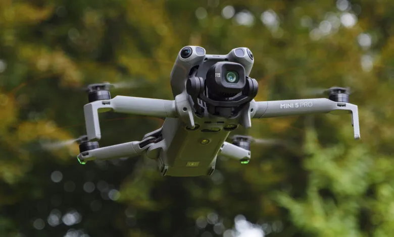 A DJI Mini 5 Pro drone in flight, showcasing its camera and propellers against a blurred green background.