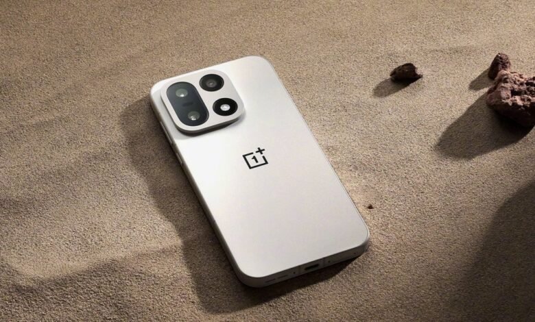 A light-colored OnePlus smartphone rests on a sandy surface with rocks.