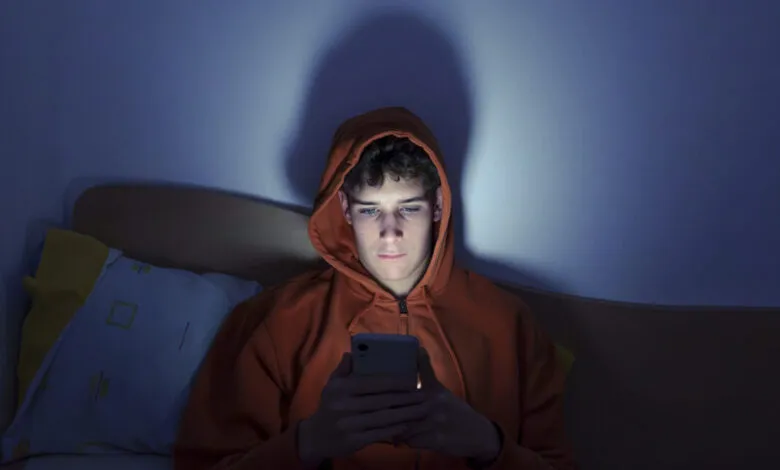 Teenager in orange hoodie uses smartphone in bed at night, illuminated by screen light.