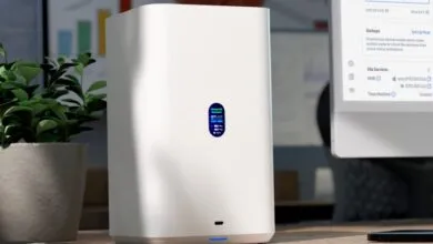 White network storage device on a desk, next to a plant and computer showing file backups and Time Machine.