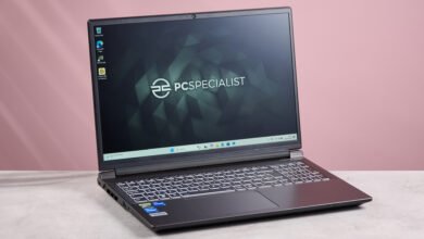 PC Specialist Defiance laptop displayed on a table against a pink background. The laptop screen shows the Windows desktop.