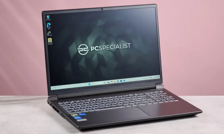 PC Specialist Defiance laptop displayed on a table against a pink background. The laptop screen shows the Windows desktop.