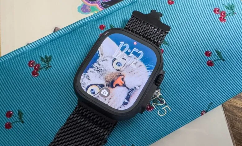 Black Apple Watch with a funny cat wallpaper on a teal cherry-patterned surface.