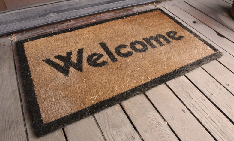 Tan welcome mat with black lettering on a wooden porch.