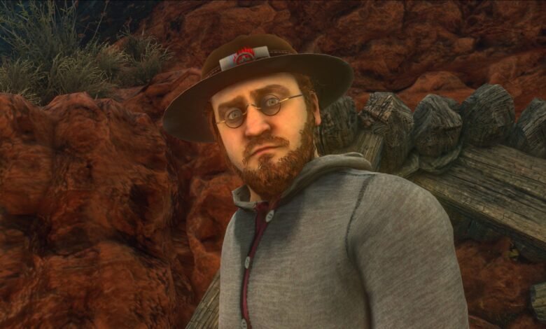 Close-up of a man in a hat and glasses, looking directly at the viewer. He has a beard and is wearing a grey hoodie.