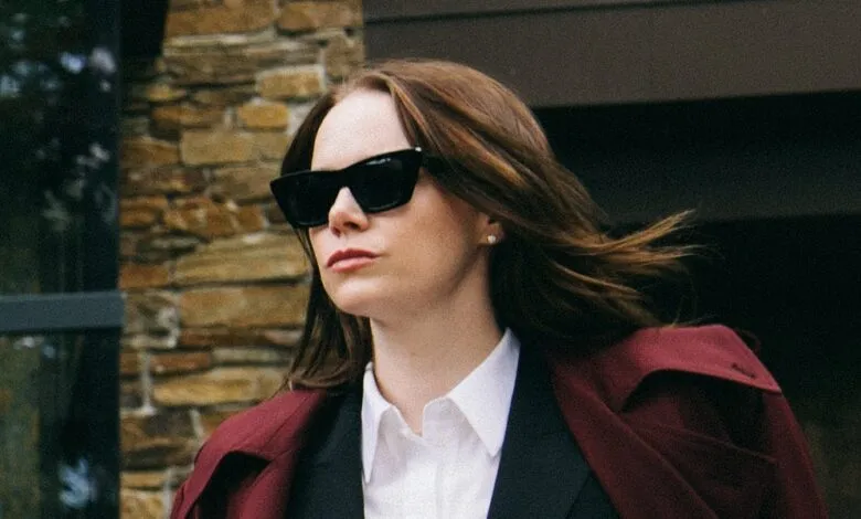 Woman with auburn hair wearing sunglasses and a maroon coat over a white shirt.