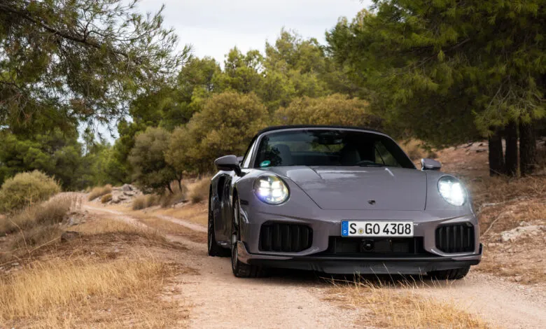 A grey Porsche 911 Turbo S convertible drives on a dirt road through a forest.