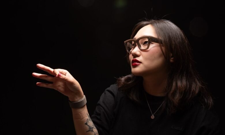 Woman wearing glasses and a black shirt, holding up three fingers against a dark background.