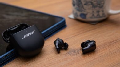 Bose QuietComfort Ultra Earbuds with charging case on a wooden surface next to a tablet and mug.