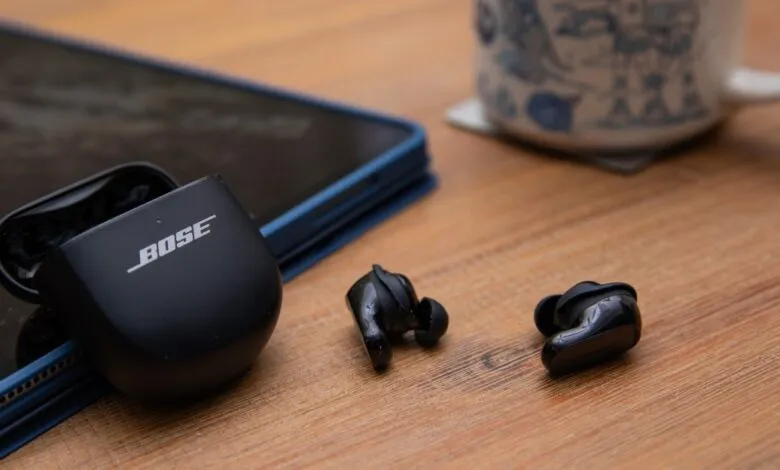 Bose QuietComfort Ultra Earbuds with charging case on a wooden surface next to a tablet and mug.