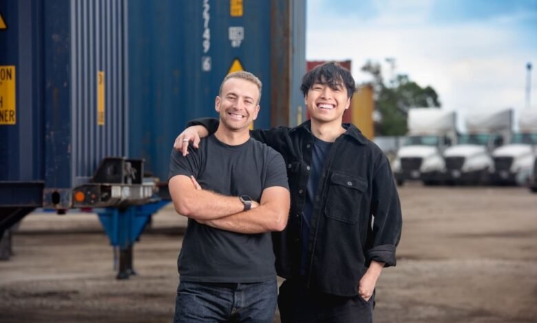 Two smiling men stand with arms around each other in front of shipping containers and trucks.