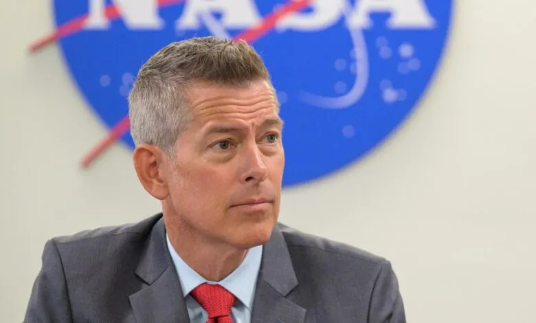 Man in a suit with a red tie looking to the side, with a NASA logo in the background.