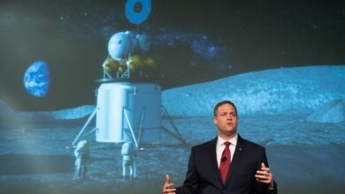Man in suit speaking in front of a projection of a lunar landing with astronauts.