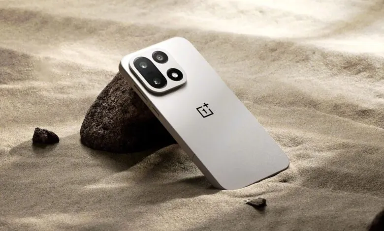A sleek, silver OnePlus smartphone rests against a dark rock on a sandy surface.