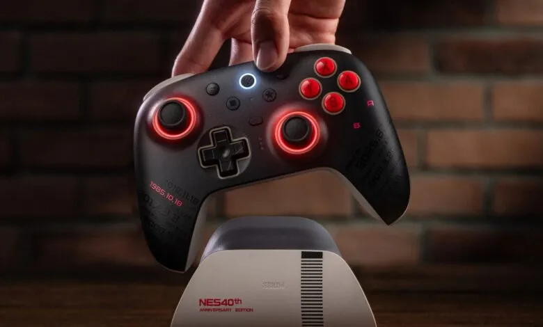 A hand holds a black 8BitDo NES 40th Anniversary Edition controller with red illuminated accents.