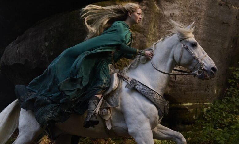 A blonde woman in a flowing green gown rides a white horse through a rocky, wooded area.