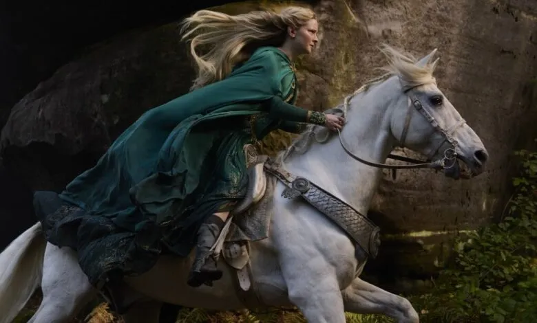 A blonde woman in a flowing green gown rides a white horse through a rocky, wooded area.
