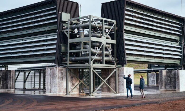 Two people stand near large industrial cooling towers with complex piping systems.