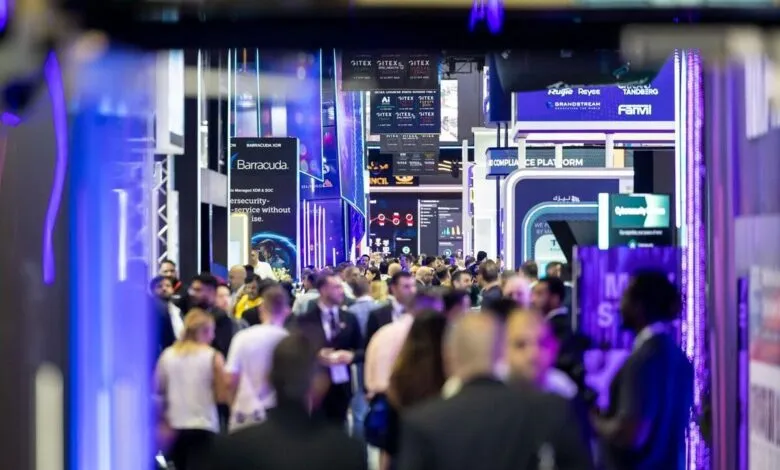 Crowd of people walking through a brightly lit exhibition hall with digital displays and company booths.