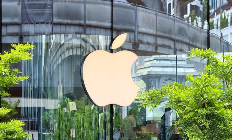 Illuminated Apple logo on a modern glass building facade with lush green trees.