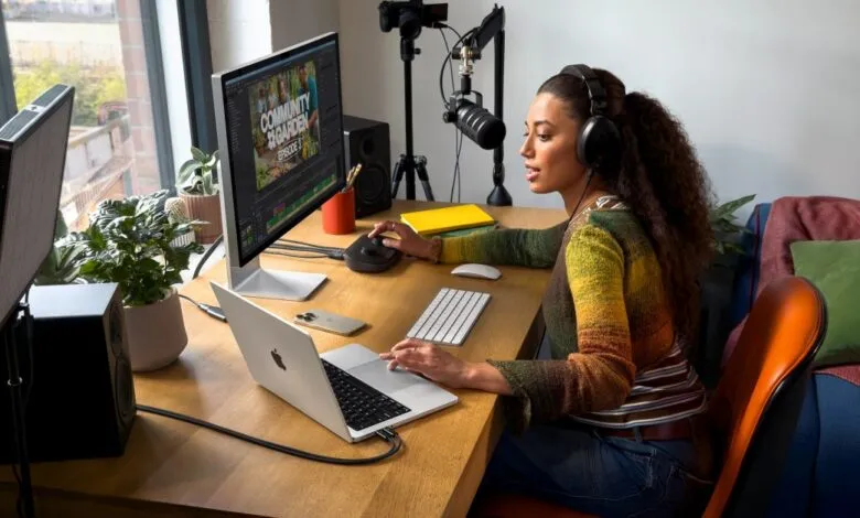 Woman editing video and recording audio at a desk with a MacBook Pro and microphone.