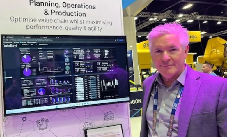 Man in a purple suit standing next to a large screen displaying production data and charts.